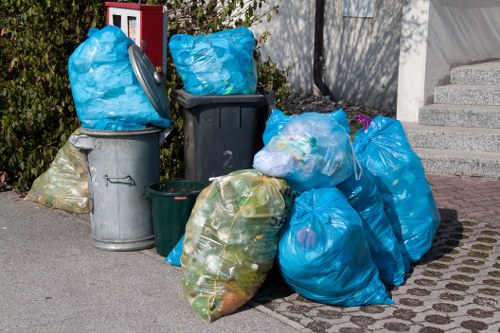 A collection of various rubbish bags and waste containers placed on a paved sidewalk in an urban environment, with some bags filled with plastic bottles and general waste, predominantly in blue and clear plastic, while another is dark green. The bags are piled next to metal and plastic bins, including a grey metal bin with a rounded lid and a blue plastic bag tied at the top. The background features a city street scene with a cement staircase on the right and some greenery on the left, possibly part of a residential or commercial area. The image reflects typical waste disposal of household rubbish, with the rubbish bags and bins awaiting collection, aligning with services provided by Garden Clearance Leytonstone in the local postcode area. The scene is well-lit, suggesting daytime, with natural light and visible textures of the plastic bags, metal, and concrete surfaces.