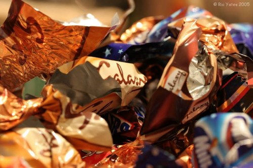 A close-up photograph of a collection of crumpled foil chocolate wrappers and candy packaging with metallic and glossy surfaces in various shades of copper, silver, blue, and red. The wrappers are piled together in a disorganized manner, occupying the foreground of the image. In the background, blurred elements suggest an indoor or outdoor environment associated with waste or rubbish collection, consistent with rubbish removal services such as those offered by Garden Clearance Leytonstone. The textures of the wrappers include crinkled, shiny, and smooth parts, reflecting light and creating highlights and shadows across the scene. The image emphasizes waste materials linked to consumer packaging, suitable for illustrating waste collection or rubbish disposal in a residential or commercial setting in Leytonstone or surrounding areas.