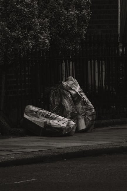 A large open area filled with a dense accumulation of mixed waste and household rubbish, including plastic bags, paper, cardboard, discarded food packaging, and other assorted debris. The waste is spr